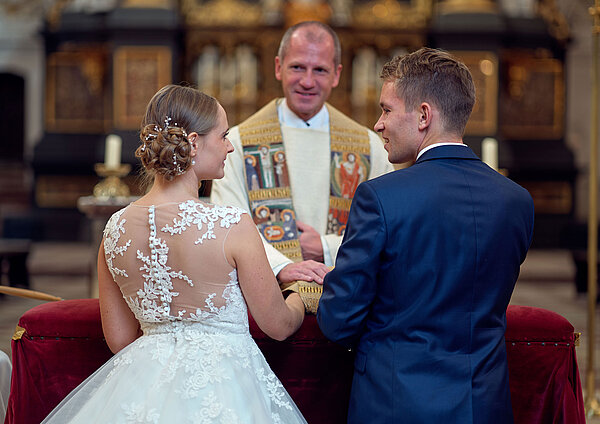 Ein junges Brautpaar erhält bei der kirchlichen Trauung den priesterlichen Segen von Dekan Roman Gerl und schaut sich dabei an.