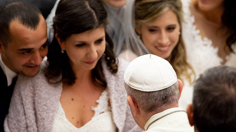 Ein frisch vermähltes Paar im Portrait, das den Segen von Papst Franziskus erhält. Im Hintergrund sind weitere Brautpaare zu erkennen. 