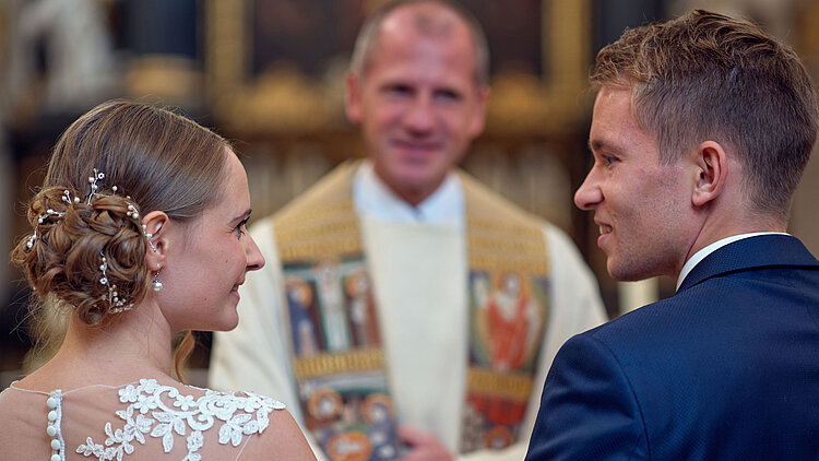 Ein junges Brautpaar erhält bei der kirchlichen Trauung den priesterlichen Segen von Dekan Roman Gerl und schaut sich dabei glücklich an.